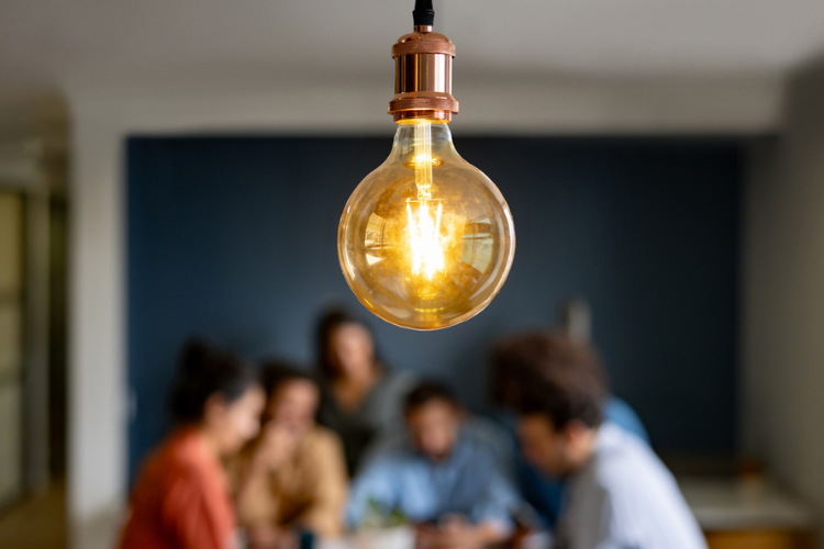 Close-up of a glowing light bulb hangs from the ceiling, with a blurred group of people collaborating at a table in the background.