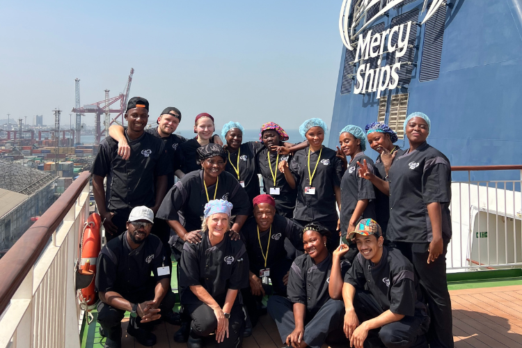 Mercy Ship volunteers pose on deck, wearing black uniforms and colorful head coverings, while behind them is a large blue ship structure with the words "Mercy Ships."